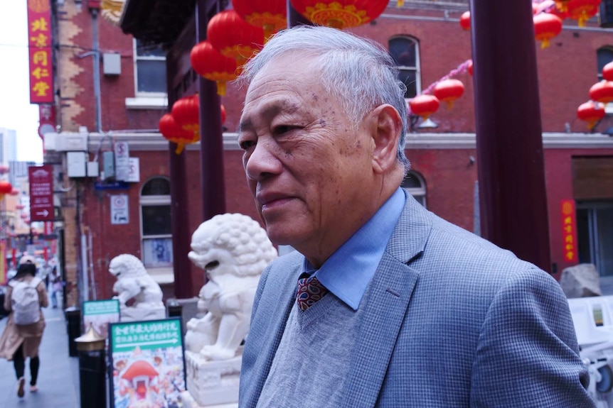 An elderly Chinese man stands in the main square of Melbourne's China town in front of red lanterns and white stone dragon