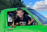 An eight-year-old boy giving the thumbs up with his dog from the window of a bright green ute.