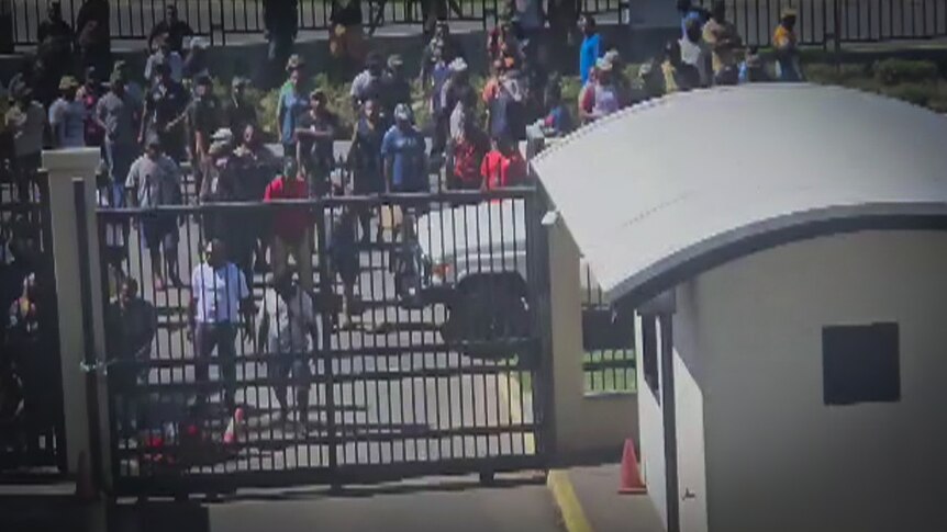 A crowd gathers outside a gate while several people try to break through the gate