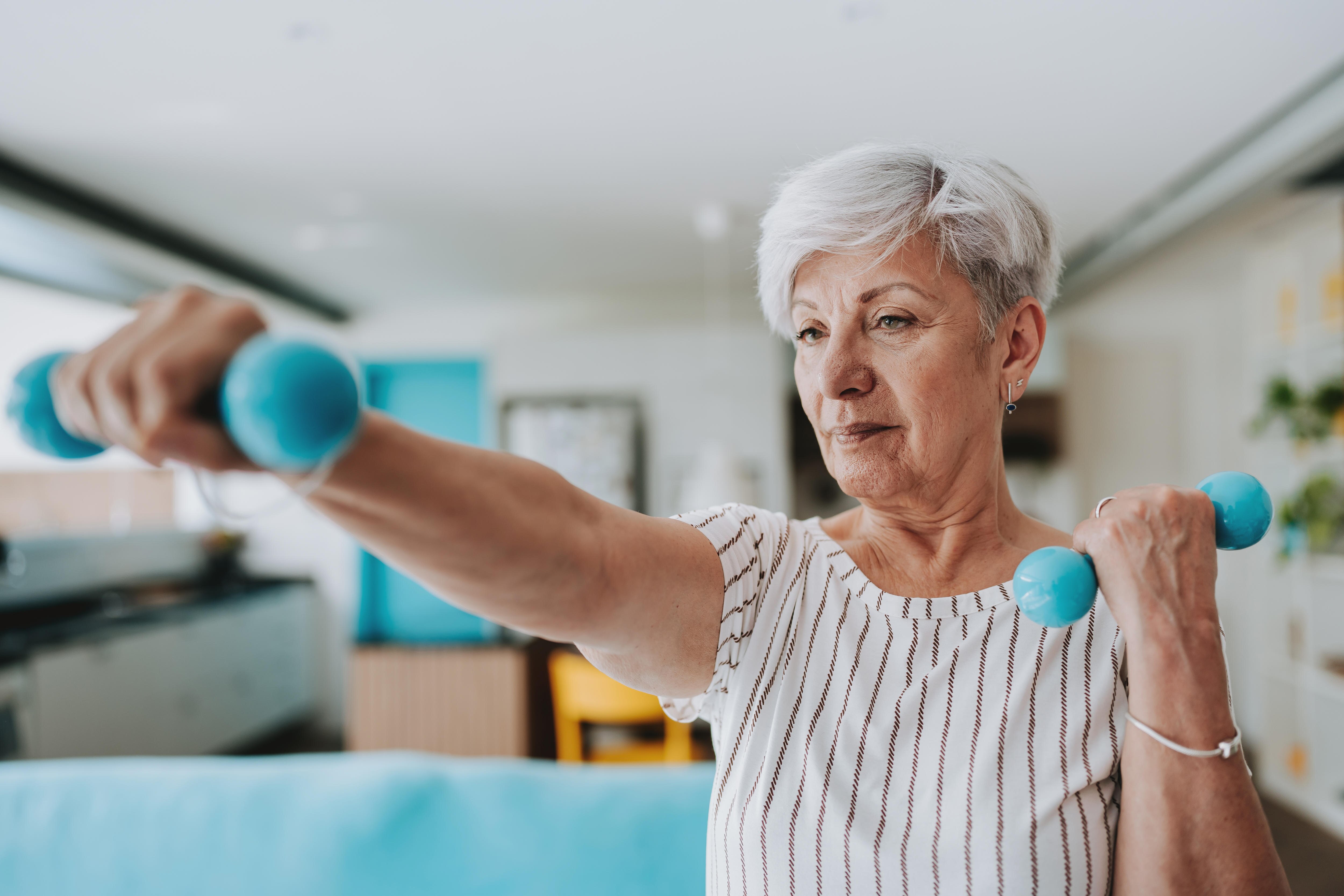 An older woman with short white hair exercising with a pair of pale blue dumb bells.