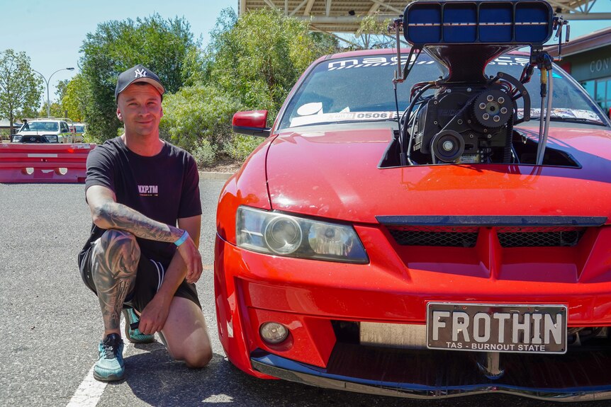 A man crouches next to a red Commodore with numberplate 'FROTHIN'