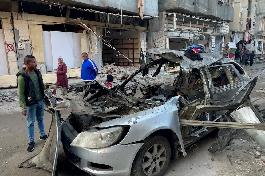 A car destroyed by an explosive device sites in the street as people look on and inspect it