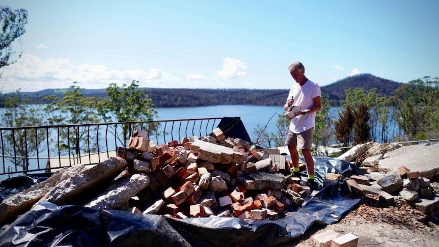 A man stands on a pile bricks while using a tool to clean one of the bricks. Behind him is a view of Lake Conjola.