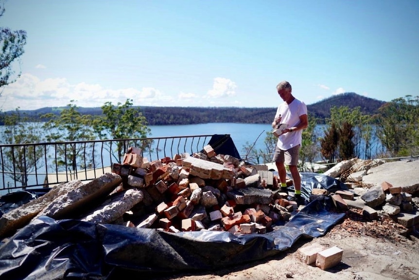 A man stands on a pile of building rubble using a tool to clean one of the bricks. Behind him is a lake and blackened hills