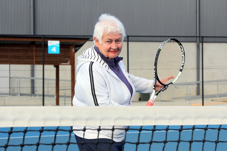 Elderly woman stands on a tennis court with her tennis racket in hand, ready to hit the ball over the net.