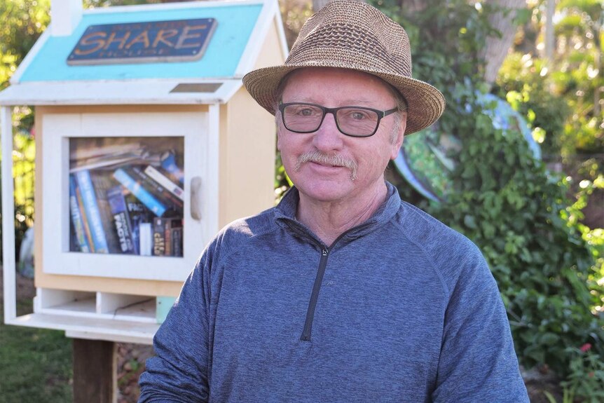 Bill Ganon wears a brown hat, glasses and smiles at the camera. Little library and greenery in the background.