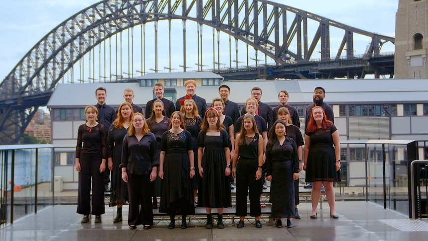 A group of young adult singers perform in 3 lines of different heights. The Sydney Harbour Bridge is behind them.