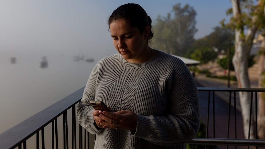 A young woman, with a misty river in the background, stares down at her phone sadly.