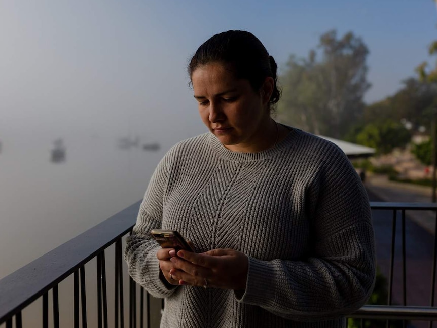 A young woman, with a misty river in the background, stares down at her phone sadly.