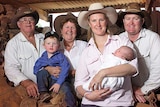 A family of 6, including two young children stand in a farm shed wearing cowboy hats.