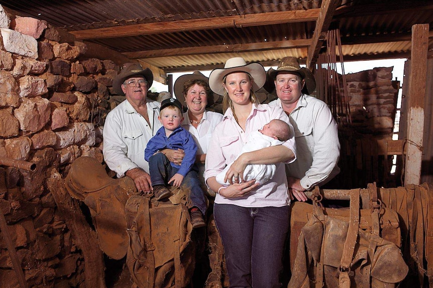 A family of 6, including two young children stand in a farm shed wearing cowboy hats.