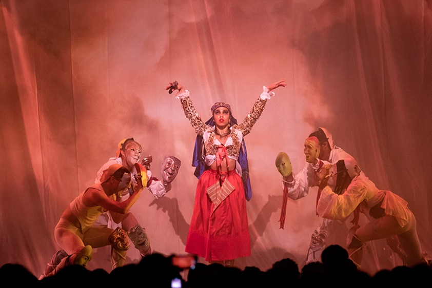FKA twigs stands on stage with her hands held up, surrounded by four masked performers