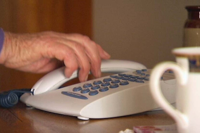 The hand of an elderly woman reaches to answer a landline phone.