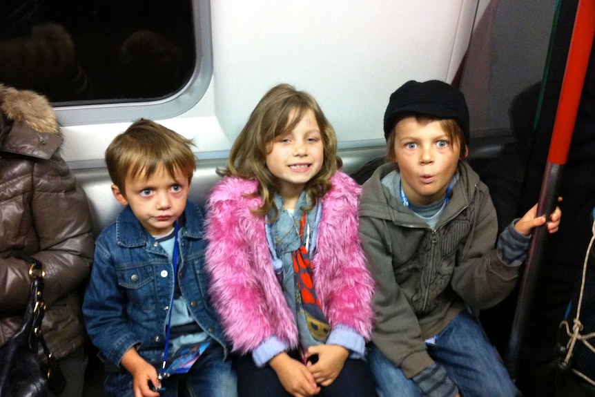 A girl dressed in pink with a beanie on, sits in front of her two brothers who are wearing hats