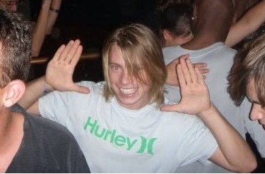 Josh wearing white shirt, smiling with hands to face and long hair.
