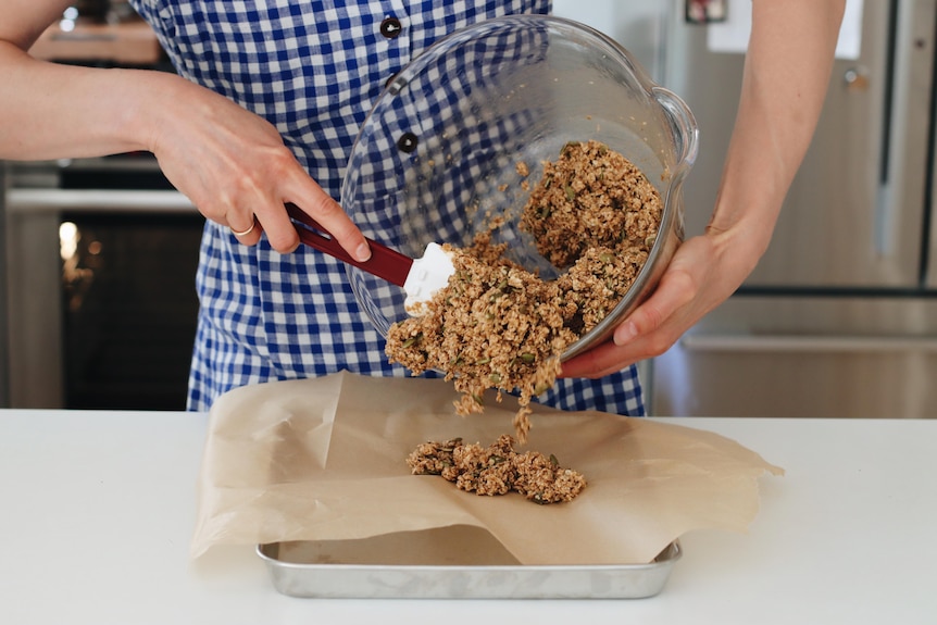 A woman in a blue gingham apron tips granola mix from a clear bowl onto a paper-lined tray.