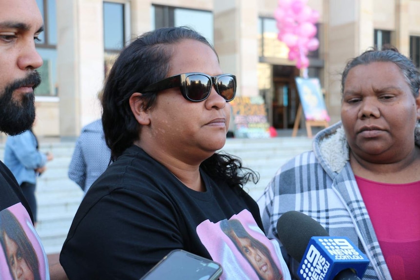 Samantha Wilson wearing black sunglasses speaking to the media, with microphones.