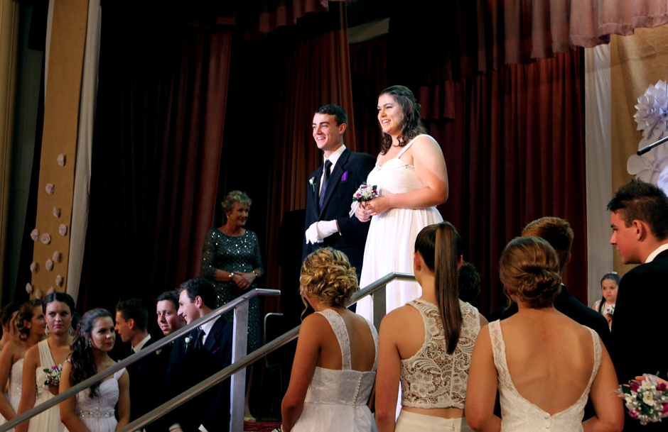 A young woman in a white dress on the arm of a suited young man, both on stage at the top of a flight of stairs.