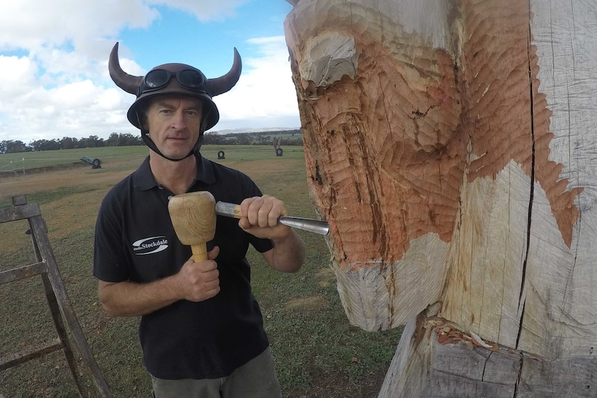 A man wearing a horned viking hat holds a hammer and chisel carving a horse head chess piece from a tree trunk.