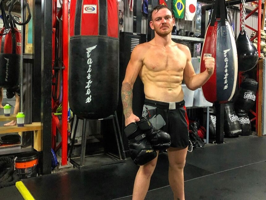 A very muscular man flexing his abs infront of a boxing bag. 
