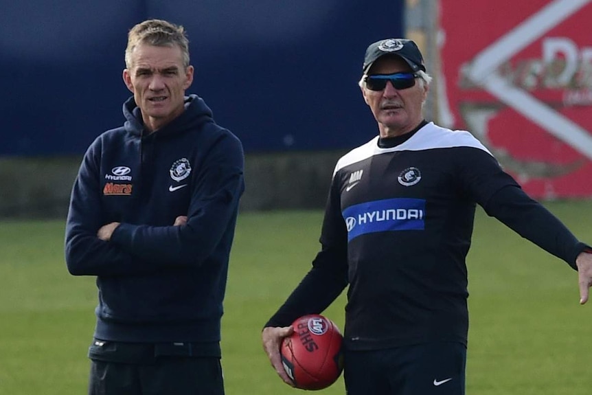 An AFL assistant coach stands with arms folded, while the senior coach holds a football at training.