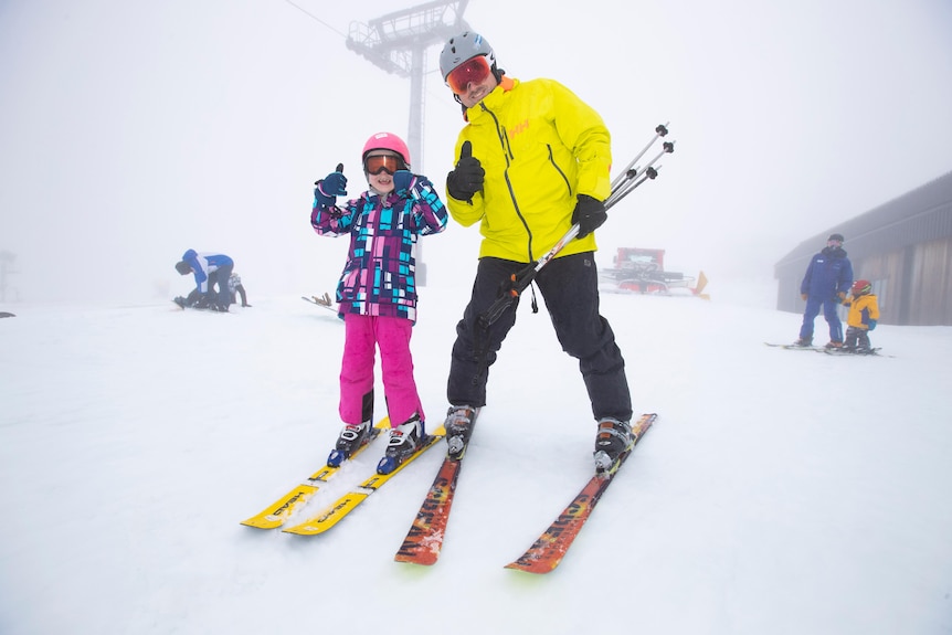 A girl and man giving the thumbs up as they ski down a slope at the snow