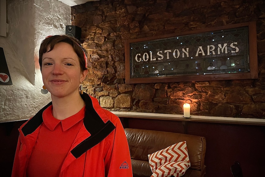 A young woman with close cropped brown hair stands next to a sign reading "Colston Arms" 