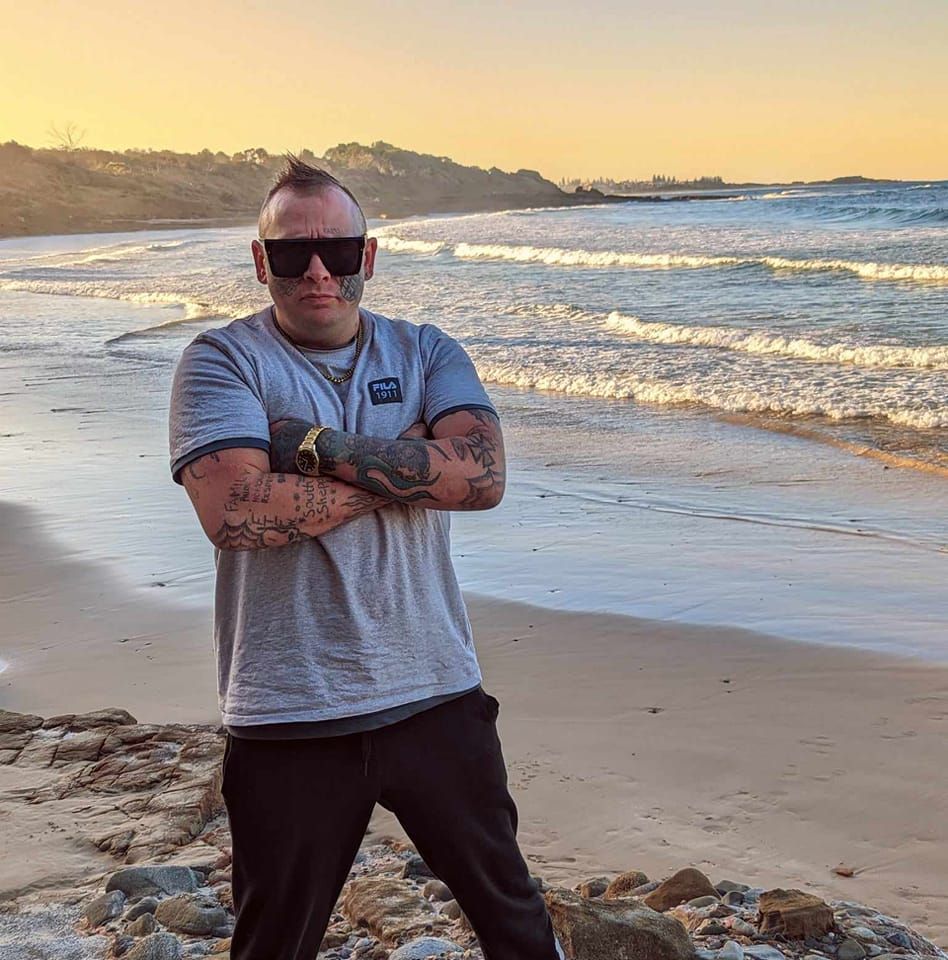 A man with sunglasses stands on the beach with his arms folded. 