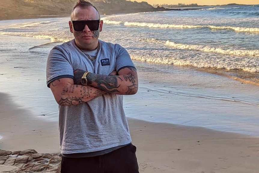 A heavily tattooed man wearing dark sunglasses stands with his arms folded on a beach.