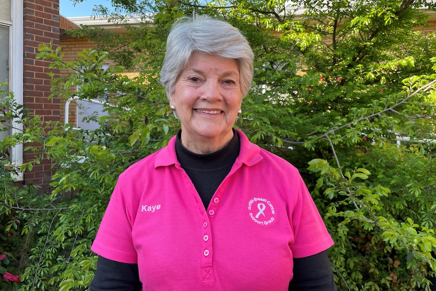 A woman with short grey hair, wearing a hot pink shirt and a black undershirt, standing in front of a tree.