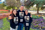 A man and two women in a park wearing identical black hoodies with a petition QR code and photo on it