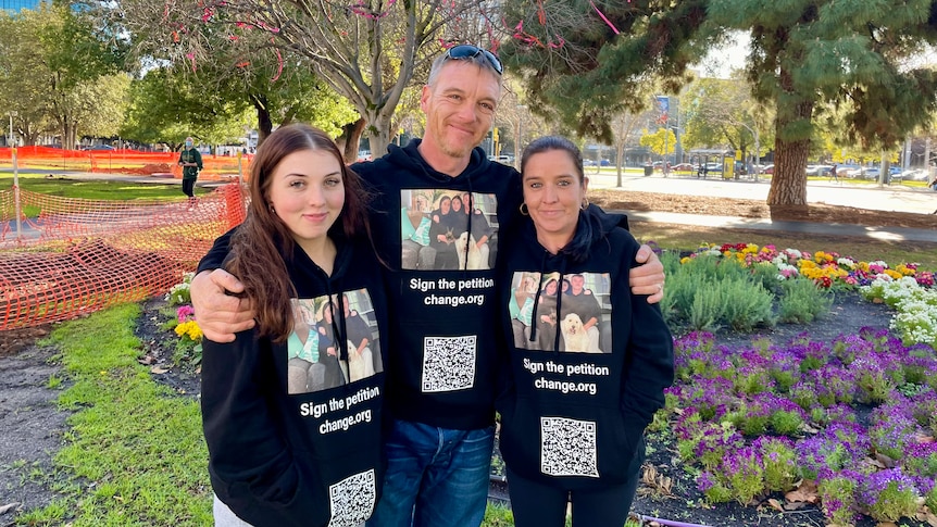 A man and two women in a park wearing identical black hoodies with a petition QR code and photo on it