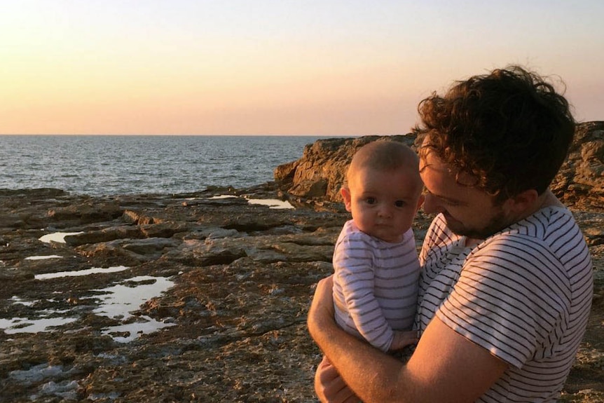 A man stands cradling a baby on some rocks in front of a seaside sunset.