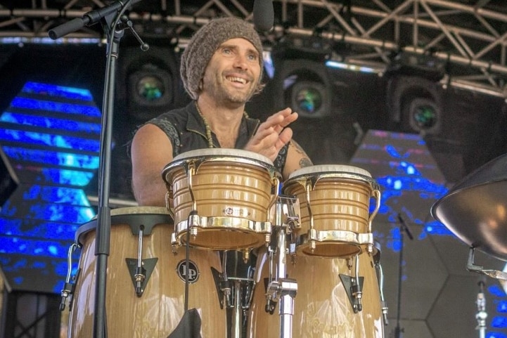 A man smiles as he plays a set of hand drums on a stage.
