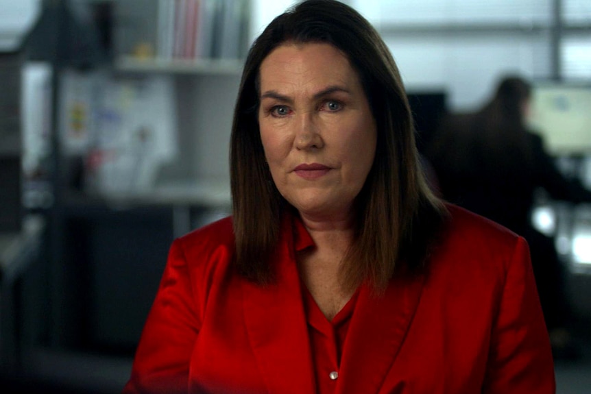 A woman wearing a red jacket sits in an office. She is looking at the camera with a neutral expression.
