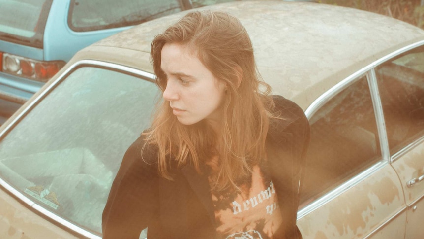 Julien wears a black jacket and sits against a brown car looking mournfully to the left.