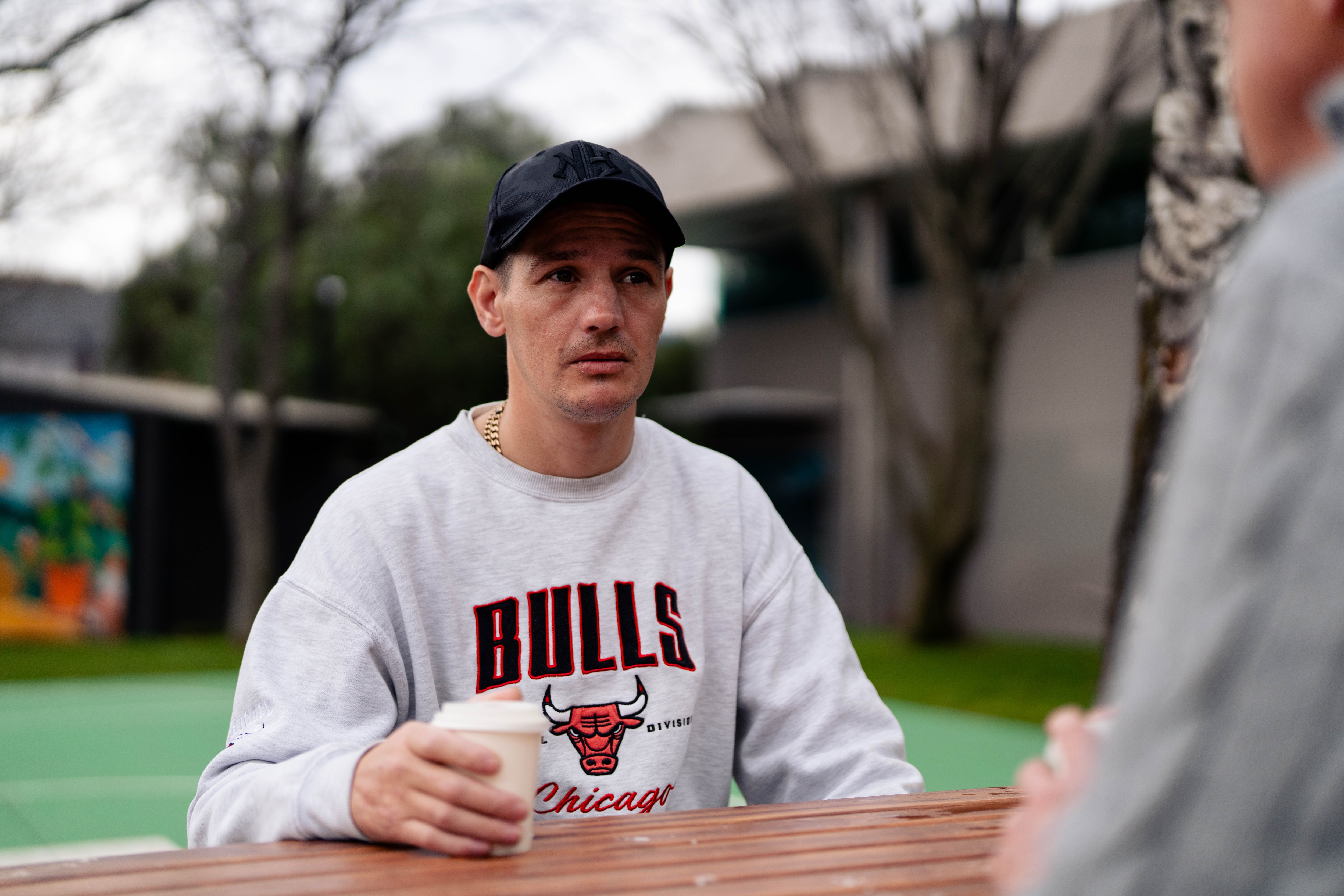 Image taken over Brendan's shoulder, looking to Shannon, wearing a grey jumper and a hat, sitting at a table, concerned.