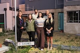 A group of people look solemn as they stand next to a mangled street sign and half-finished house in an overgrown yard.