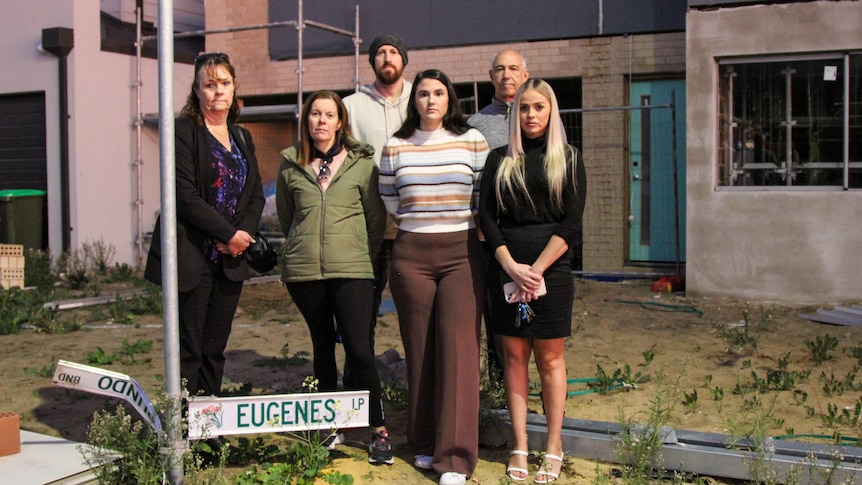 A group of people look solemn as they stand next to a mangled street sign and half-finished house in an overgrown yard.