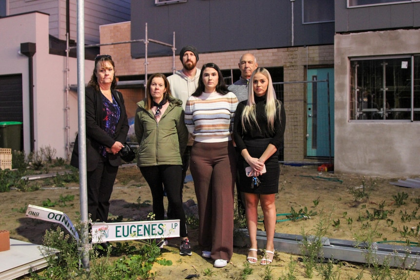 A group of people look solemn as they stand next to a mangled street sign and half-finished house in an overgrown yard.