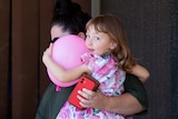 Ellie Smith carries her daughter Cleo out of a house in Carnarvon, with Cleo looking at the camera holding a pink balloon.