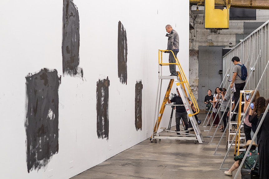 Mike Parr stands on stepladder while roughly painting black squares on a white gallery wall. Onlookers watch and take photos.