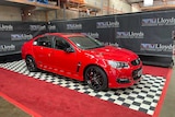 A red Holden Commodore, on a checkerboard floor, in front of banners that read 'Lloyd's auctioneers and valuers'