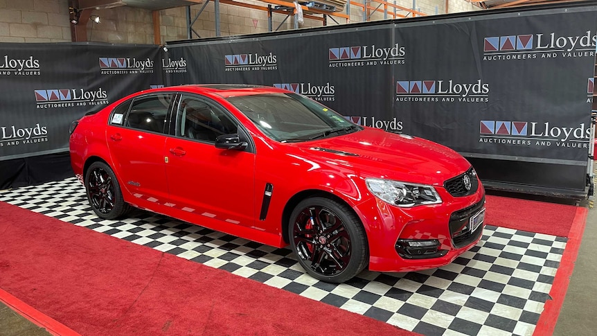 A red Holden Commodore, on a checkerboard floor, in front of banners that read 'Lloyd's auctioneers and valuers'
