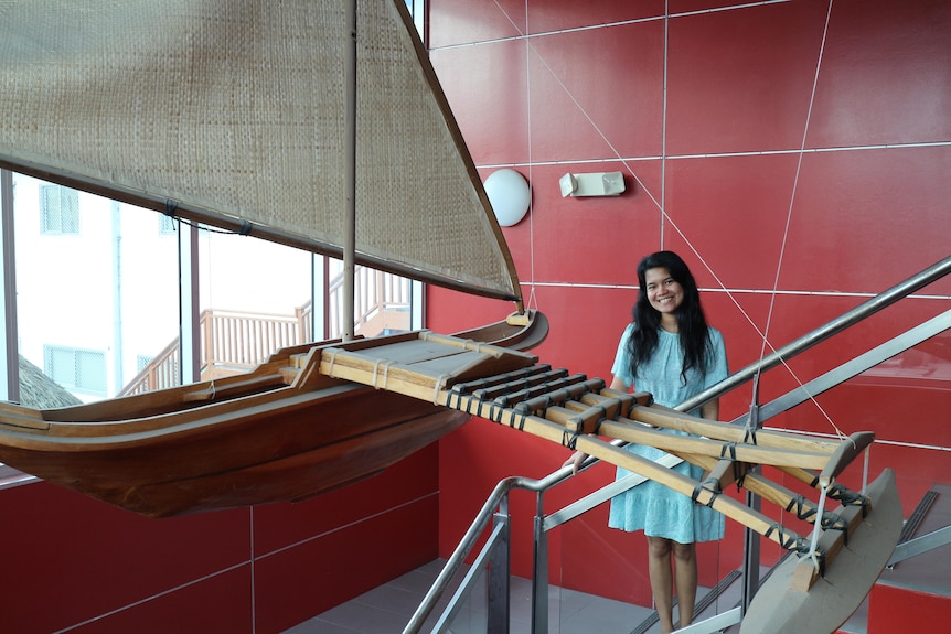 A smiling young woman with long dark hair wearing a light blue dress stands on stairs behind a model outrigger canoe