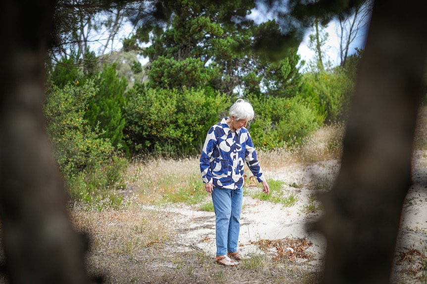 An elderly woman looks down at a fresh pile of sand in a cemetery.
