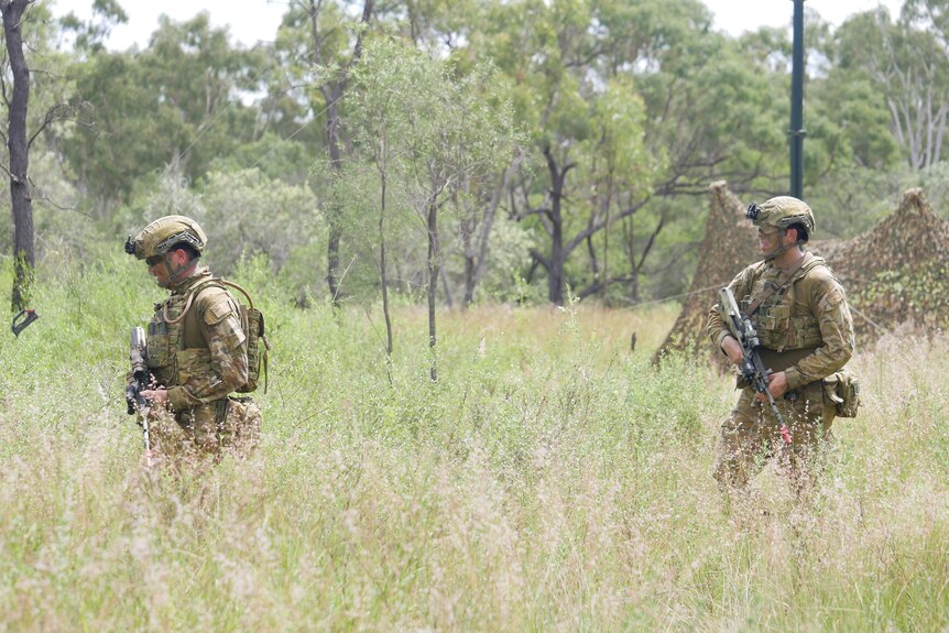 Two soldiers in camouflage walk through bushland, holding firearms.