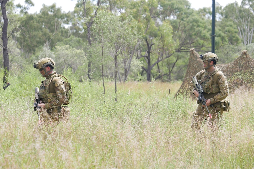 Two soldiers in camouflage walk through bushland, holding firearms.
