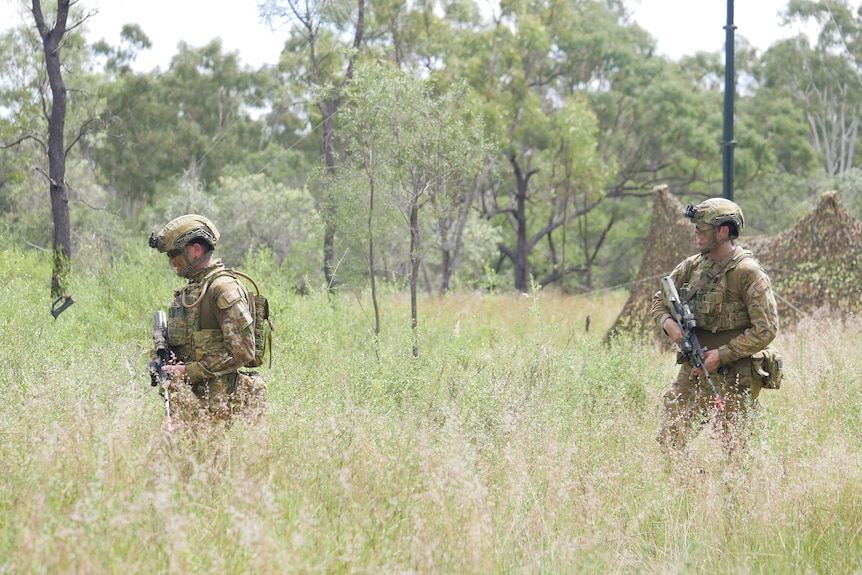 A soldier in camouflage walks through bushland holding a firearm