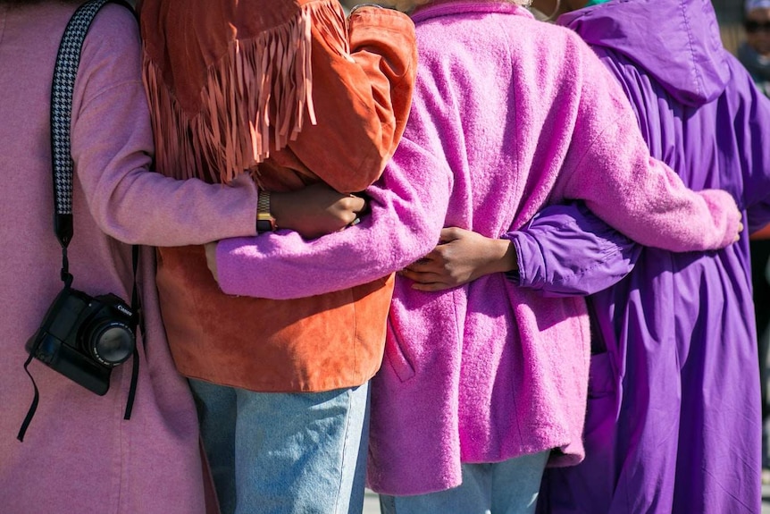 In a view of their backs, four women hug each other standing in a line. All wear bright coloured clothes.